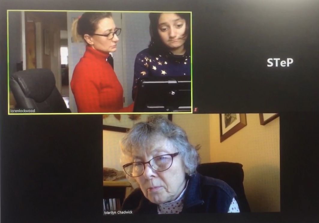 Three people in a video call. Two women stand together, one in red, the other in blue. A third woman in glasses speaks.