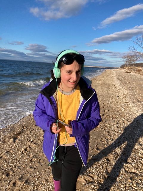 Girl with headphones and purple jacket walks on a beach. Ocean and blue sky background.