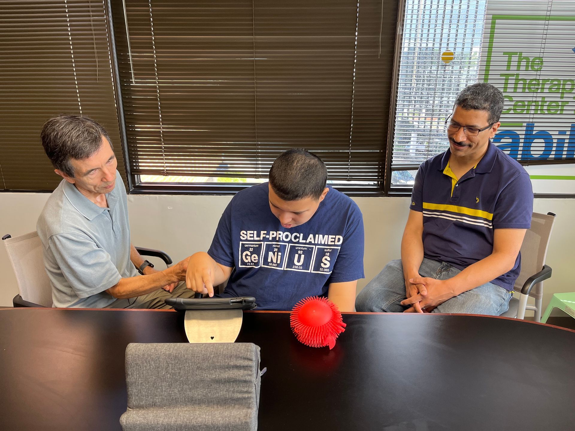 Three men at a table, looking at a tablet. Two point at the screen, one smiles. Indoors, sunny.