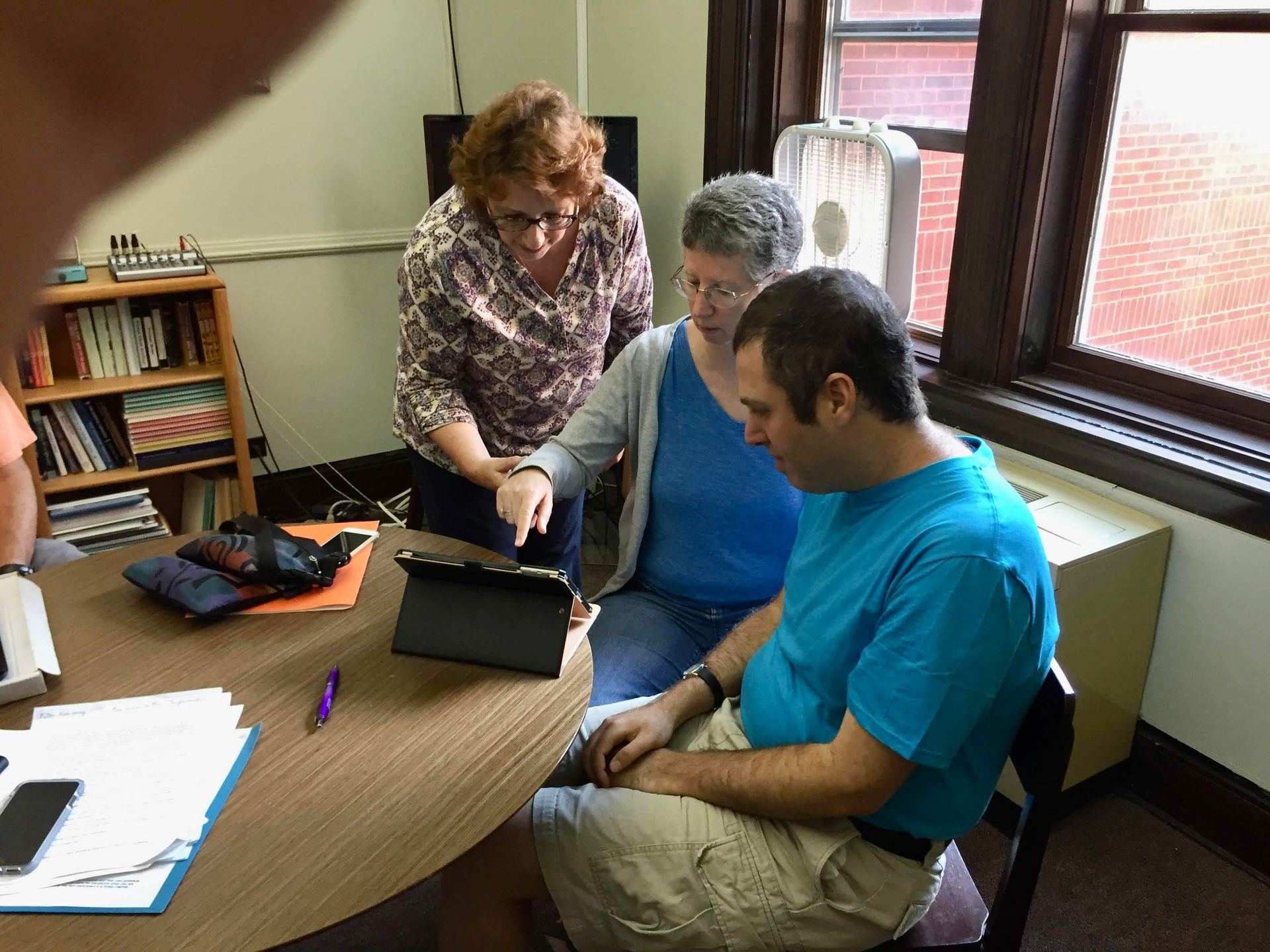 Three people at a table, looking at a tablet. Two women point at the screen, one man sits.