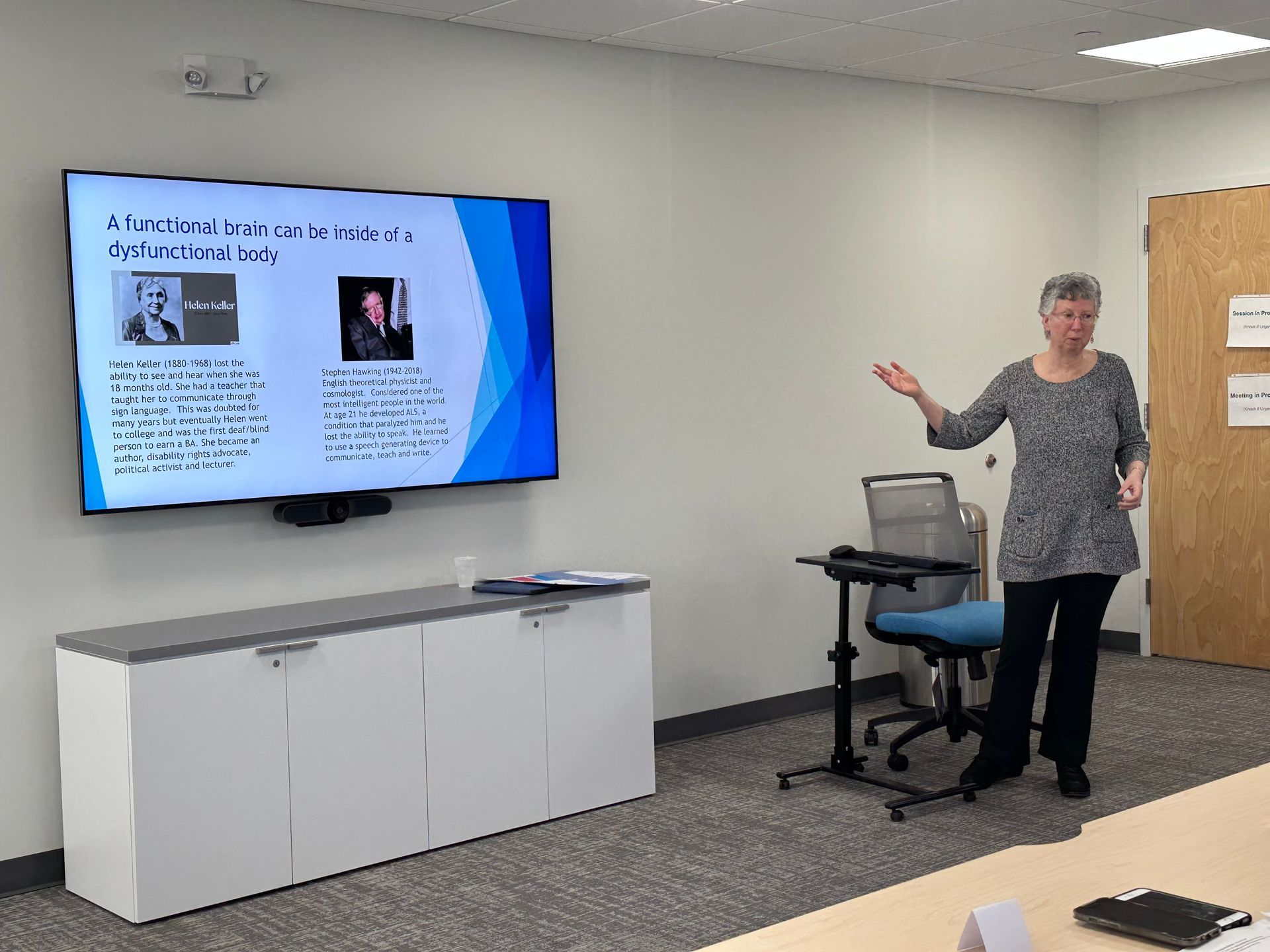 Woman presenting at a screen displaying text and photos, in a conference room.