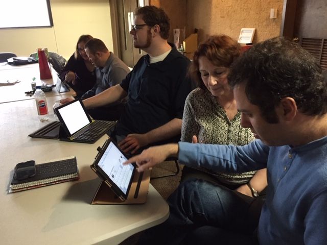 People in a meeting, some pointing at tablets on a round table.  Indoor setting, natural light.