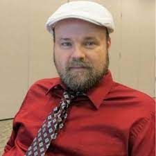Man with a beard and white hat wearing a red shirt and patterned tie.