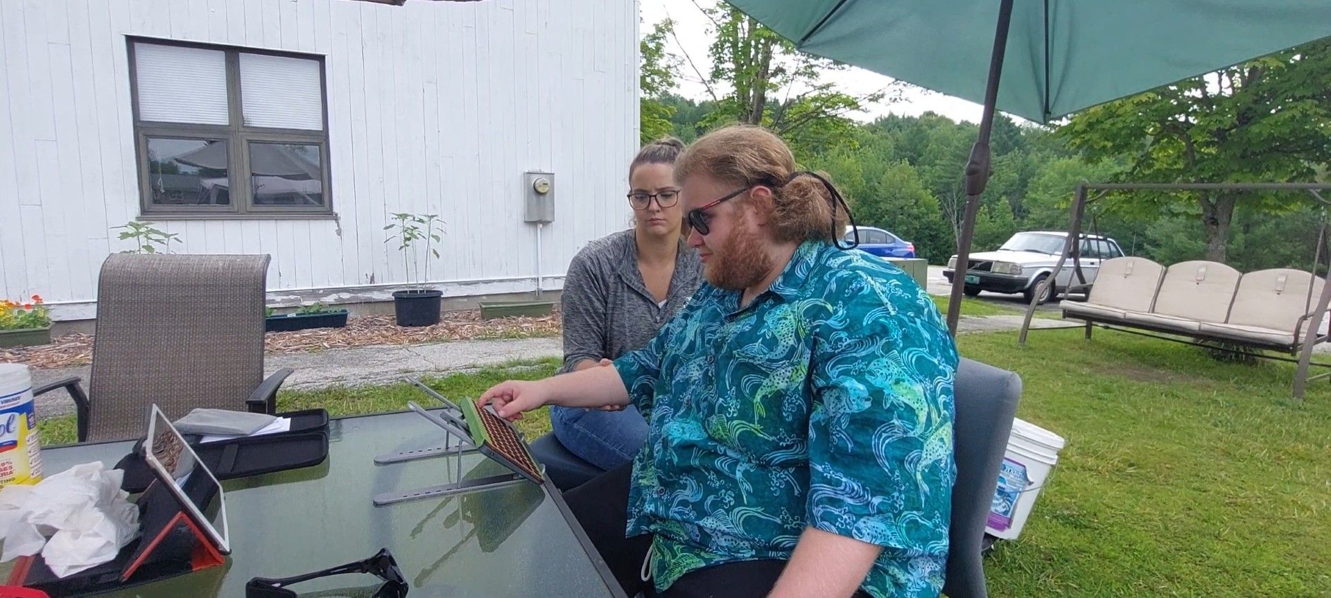 Two people outside, one pointing at a game. Man in blue shirt. Woman in glasses. Green grass, house in background.