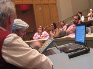 A man in a vest and a man with a tablet, laptop, and microphone in front of a classroom of people.