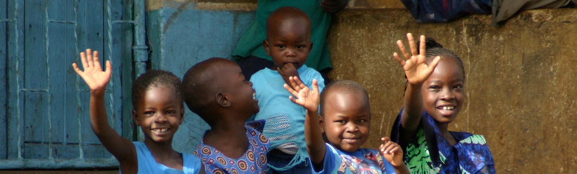 Children waving and smiling in front of a blue gate. They are wearing blue and green tops.