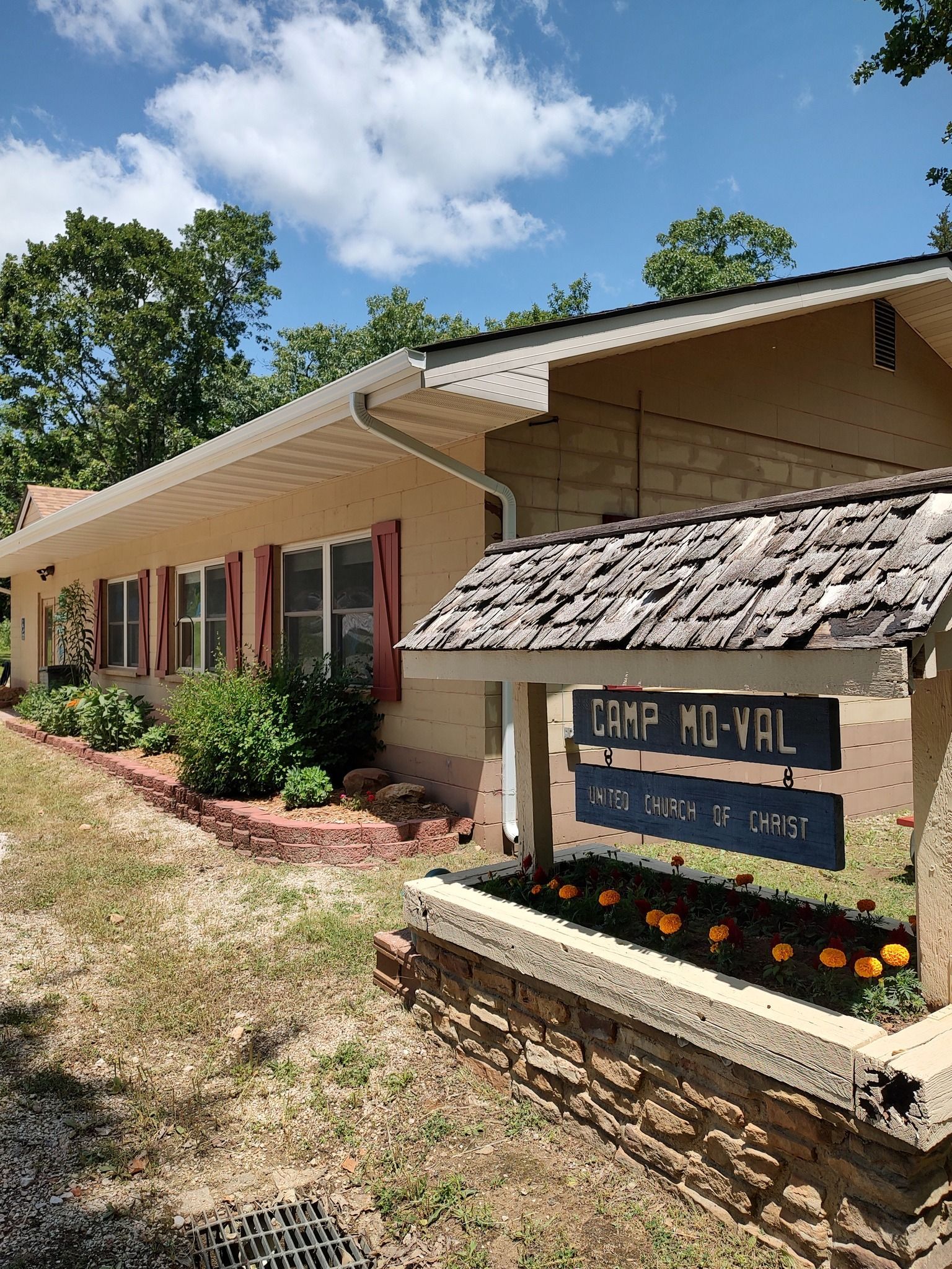Camp MoVal sign in front of a tan building with red shutters and flowers, trees, and blue sky in the background.