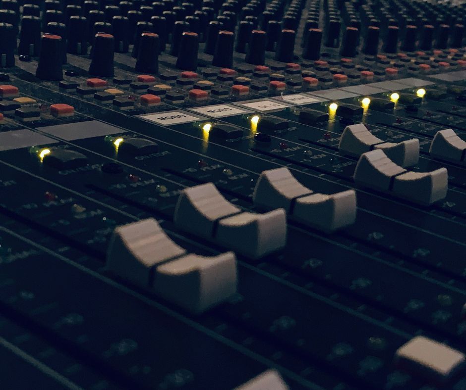 Sound mixing console with many sliders and knobs, lit up in a dark studio.