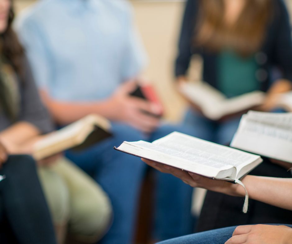 People in a circle reading open books. Focus on the open book in a person's hands. Others are blurred in background.