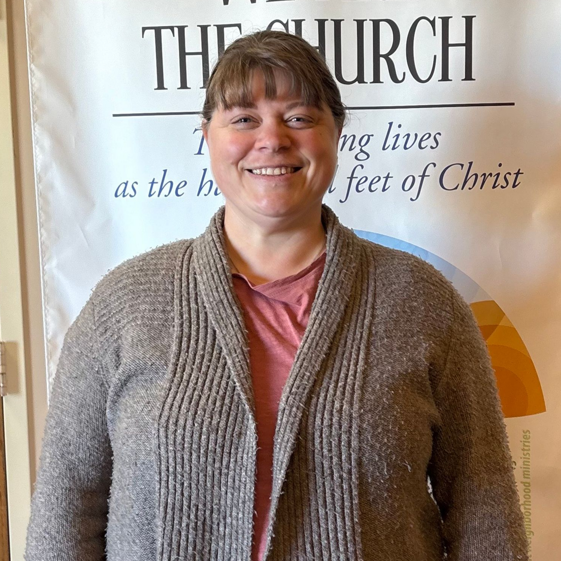 Woman smiling, wearing a gray sweater and a pink shirt, standing in front of a banner.