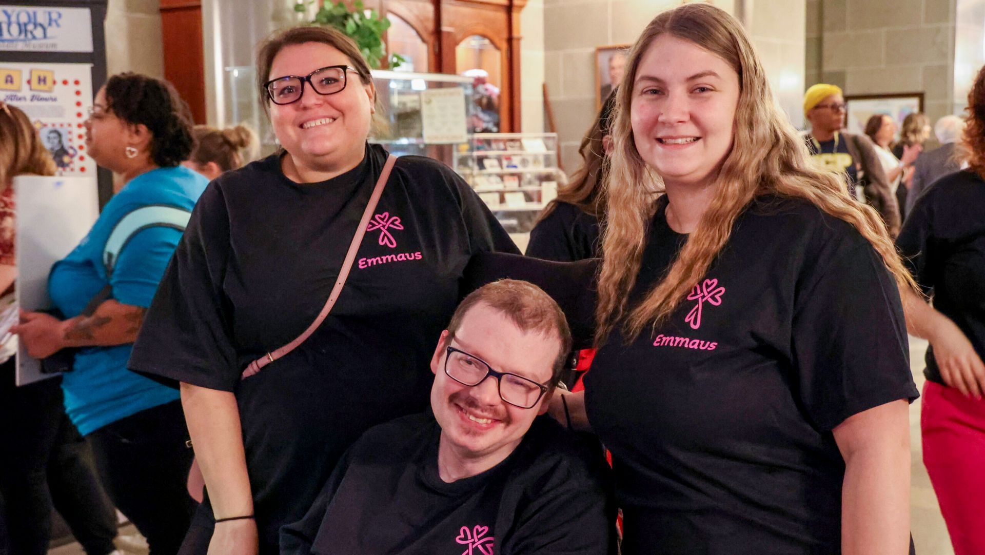 Three people smiling, wearing matching black t-shirts, posing in an indoor setting. One is in a wheelchair.