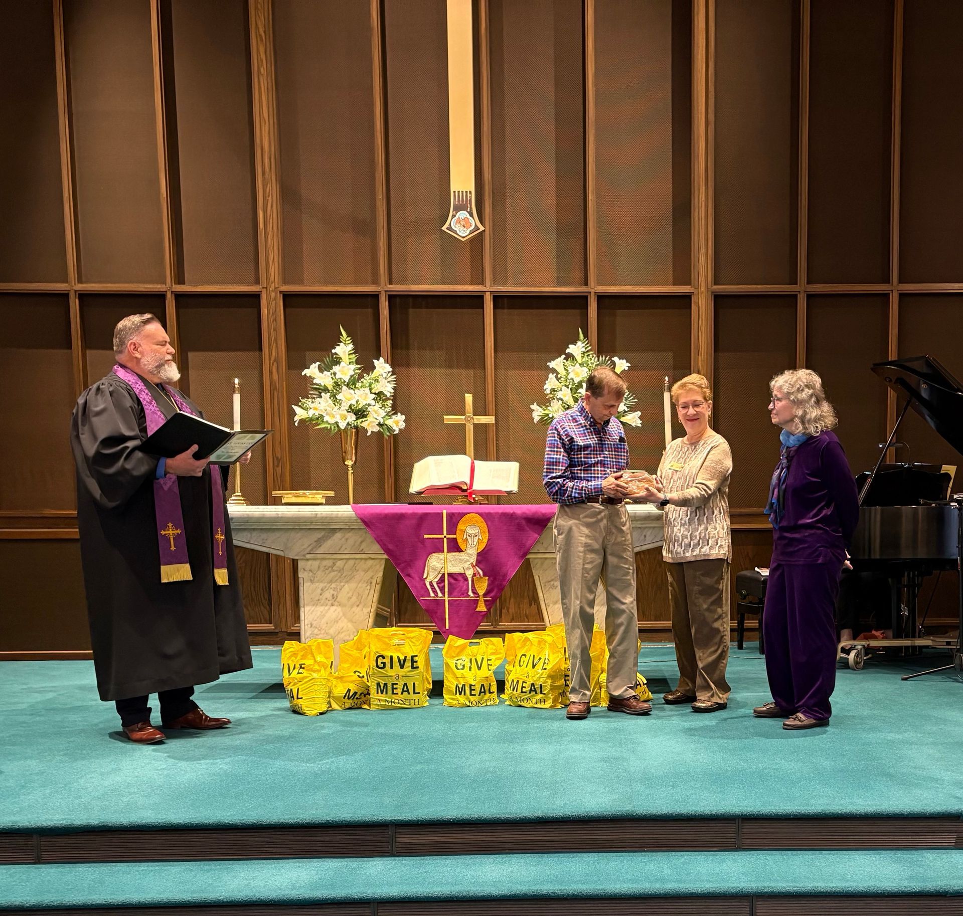 Clergy and three people at a church altar during a ceremony. Purple accents, flowers, and a piano are visible.