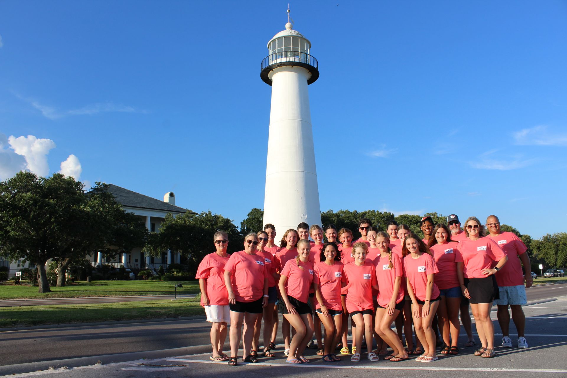 Group of people in pink shirts posing in front of a tall white lighthouse under a blue sky.