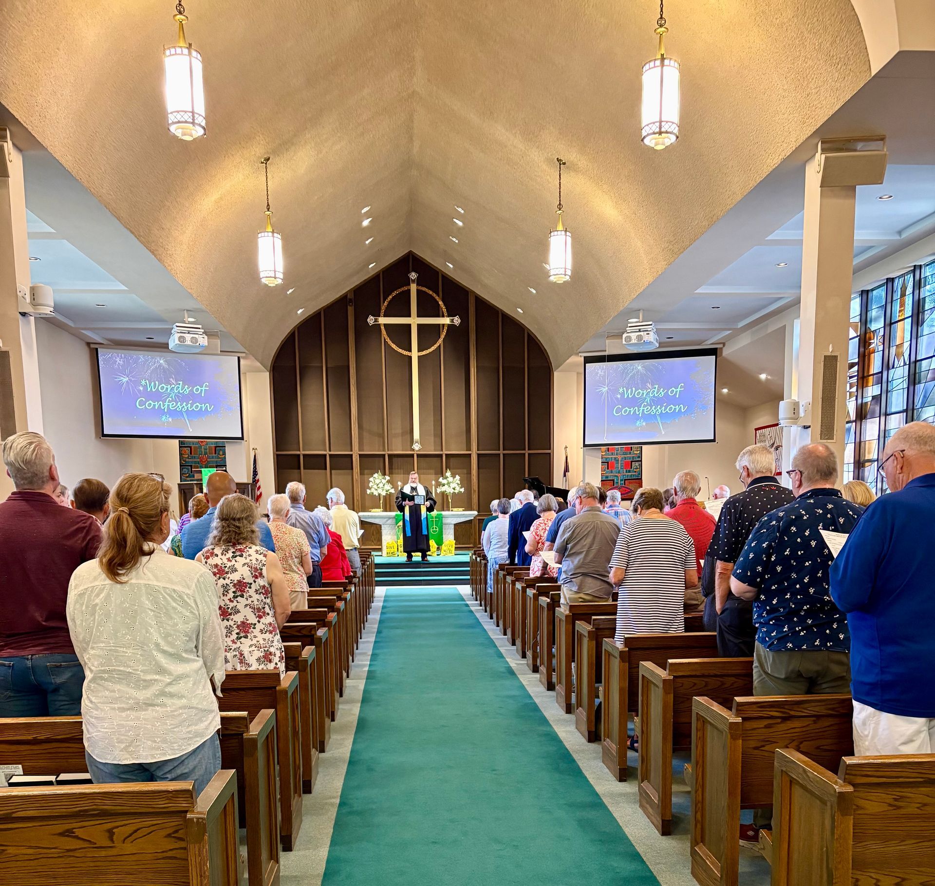 People stand in a church, facing a pastor at the altar. Pews line a teal carpet aisle. Cross visible.