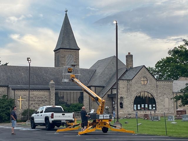 Workers on a lift installing a street light in front of a stone church. A white truck is nearby.