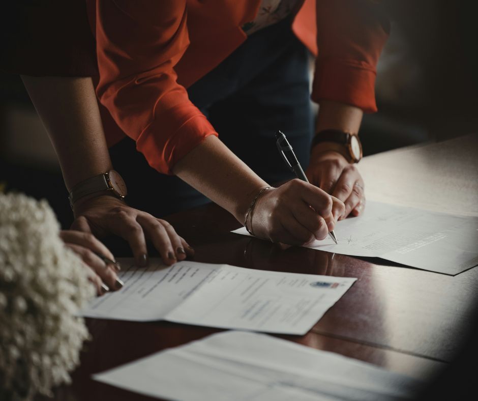 Person signing a document on a table; another person’s hands are next to the document.