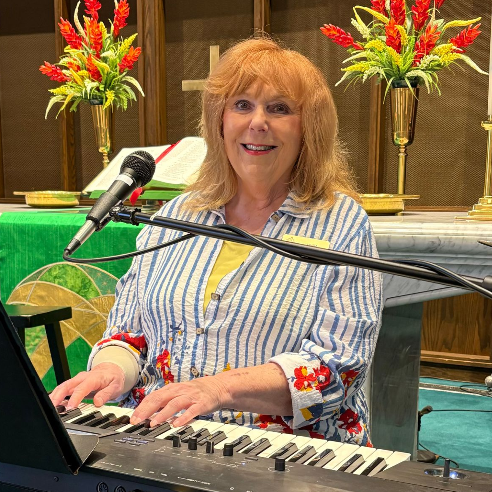 Woman playing a keyboard in a church setting, singing into a microphone. She wears a striped shirt.