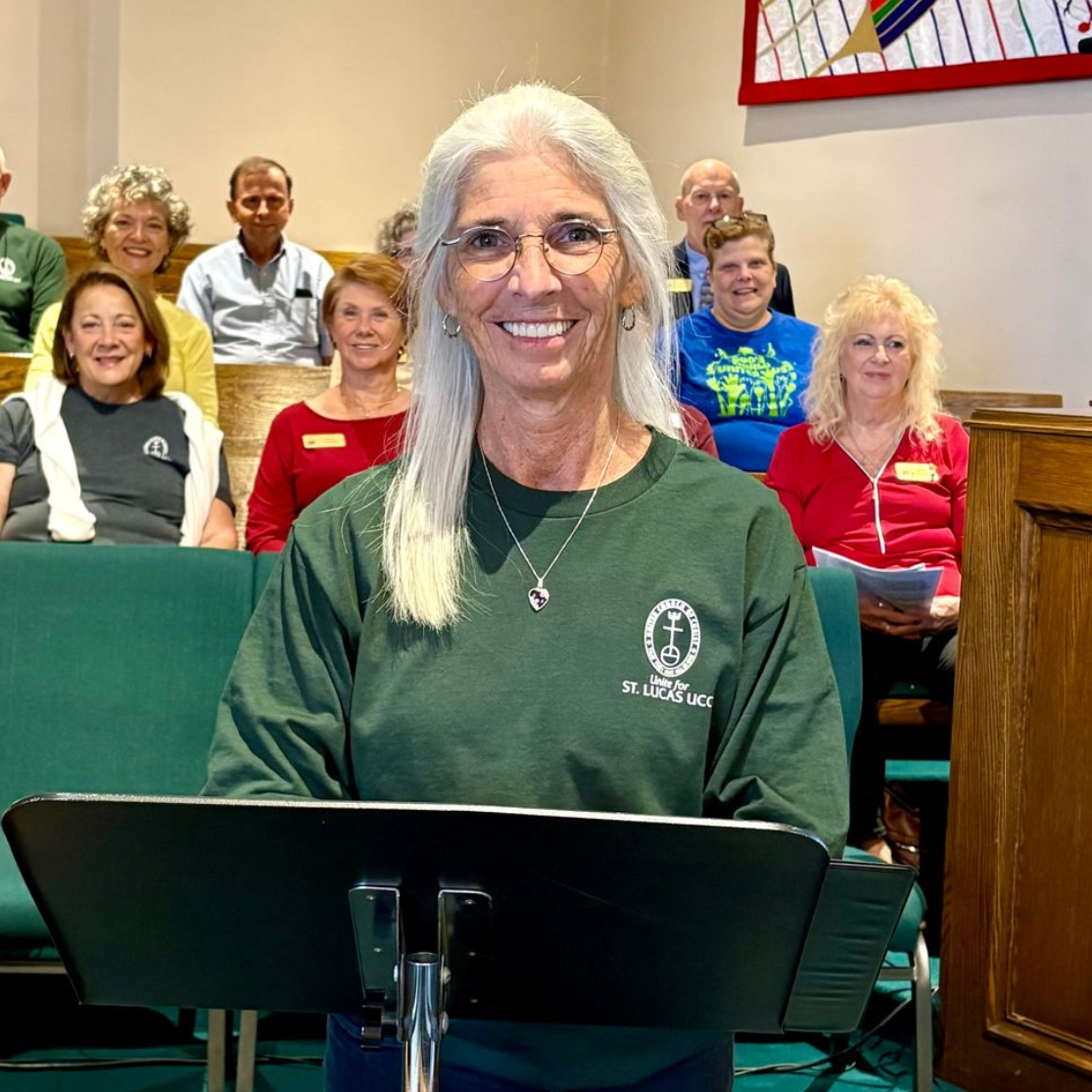 Woman in green shirt, smiling, at a podium, with a choir of people seated behind her in a church.