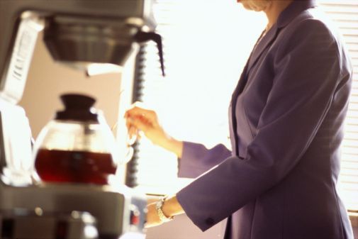 A person in a purple blazer prepares a cup of coffee using a glass carafe in a bright office setting.