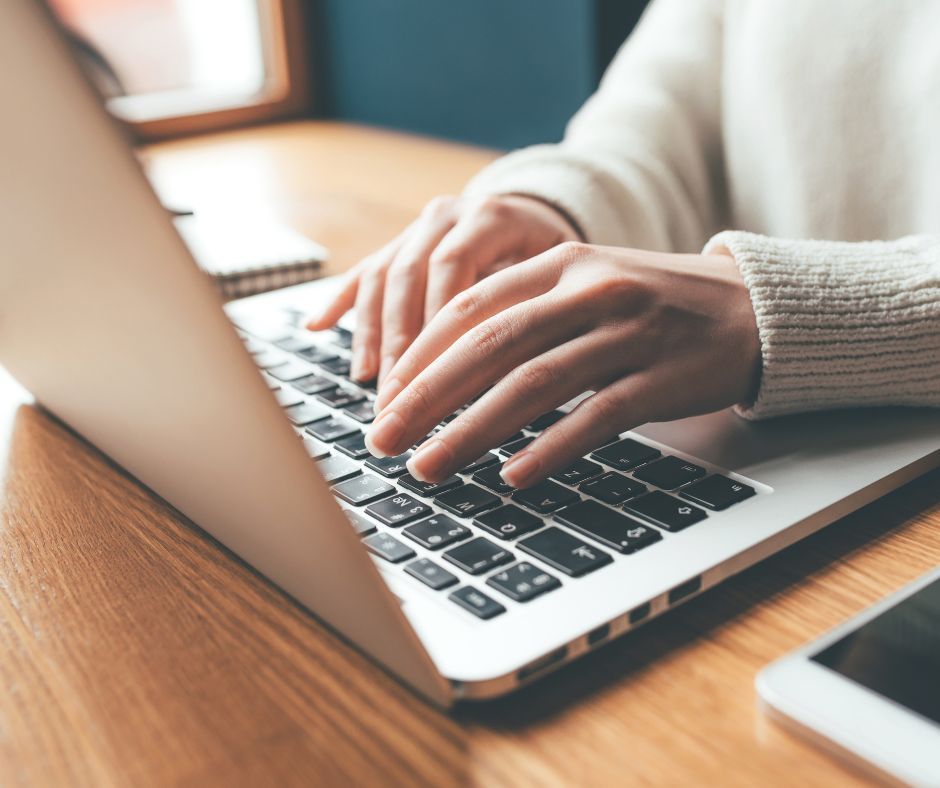 Hands typing on a laptop keyboard at a wooden desk, a smartphone visible on the right.