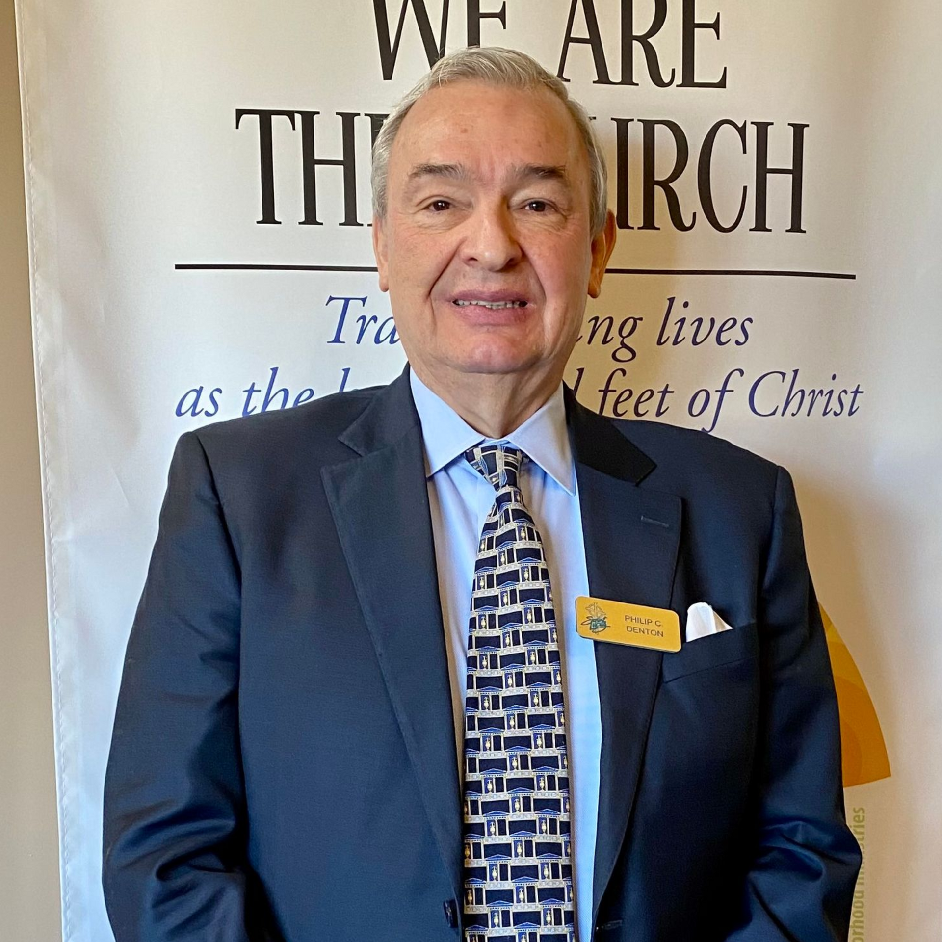 Man in a navy suit and tie, smiling, standing in front of a sign that says, 