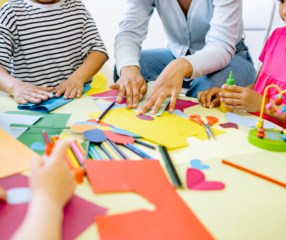 Children and adult crafting at a table with colorful paper, scissors, and glue.