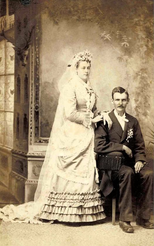Bride and groom posed, bride in white dress with train, groom in suit. Sepia tone.