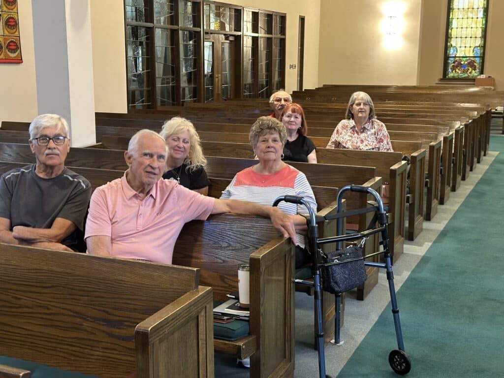 People seated in church pews, one using a walker. Interior with wood and stained glass.