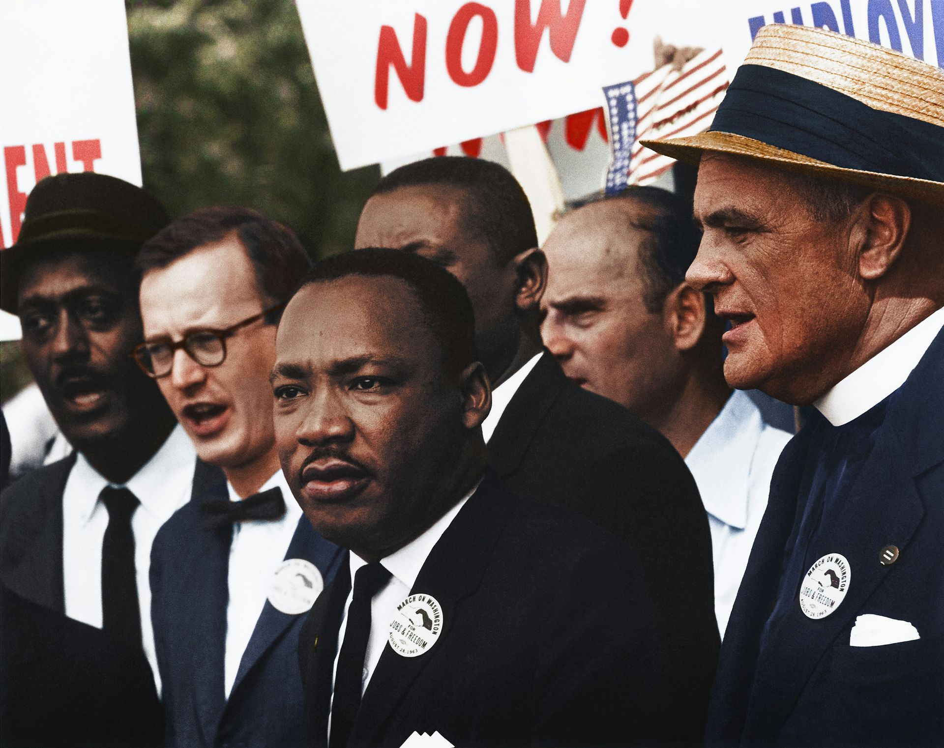 Civil rights march with Martin Luther King Jr., holding button, looking forward, signs in background.