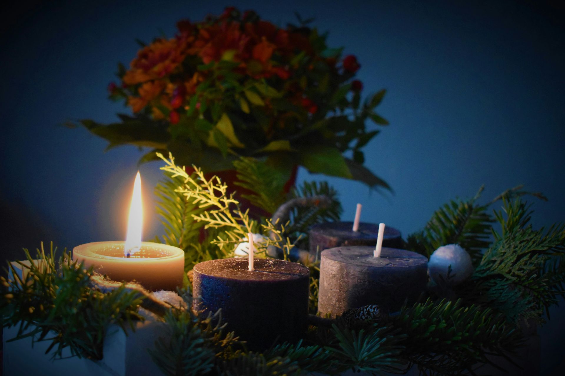 Advent wreath with lit candle, evergreen, and red flowers against a dark blue background.
