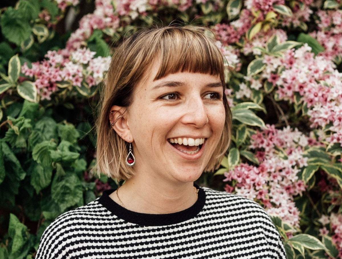 A woman is smiling in front of a bush of pink flowers.