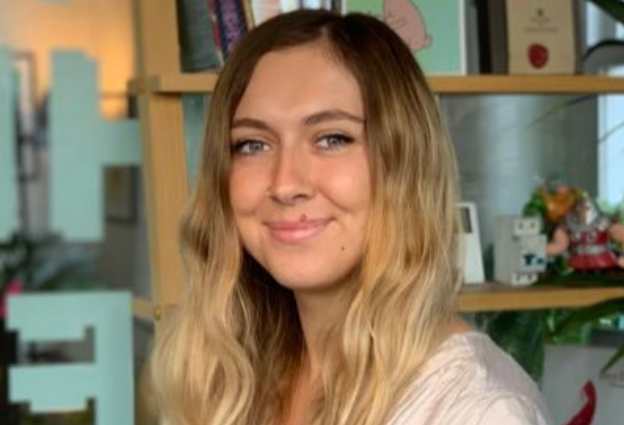 A woman with long blonde hair is smiling in front of a bookshelf.