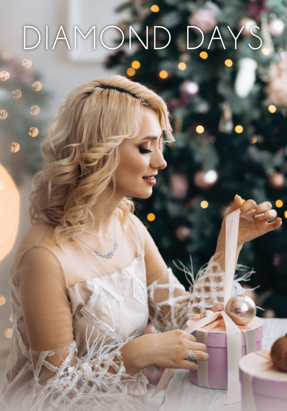 Blonde woman in a white dress opening a gift box beside festive holiday decorations and a Christmas tree