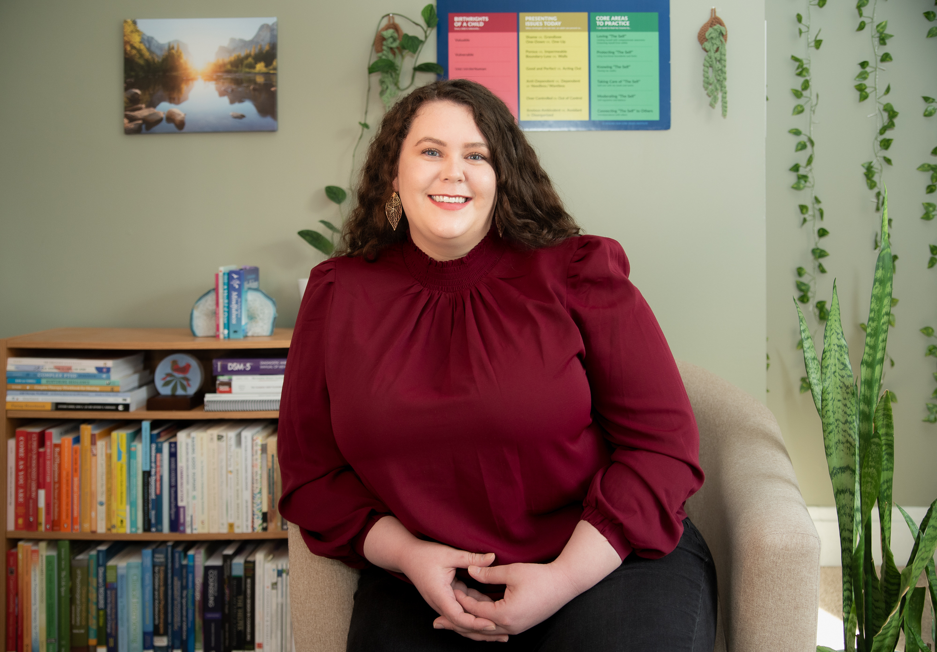 A woman in a red shirt is sitting in a chair in front of a bookshelf.