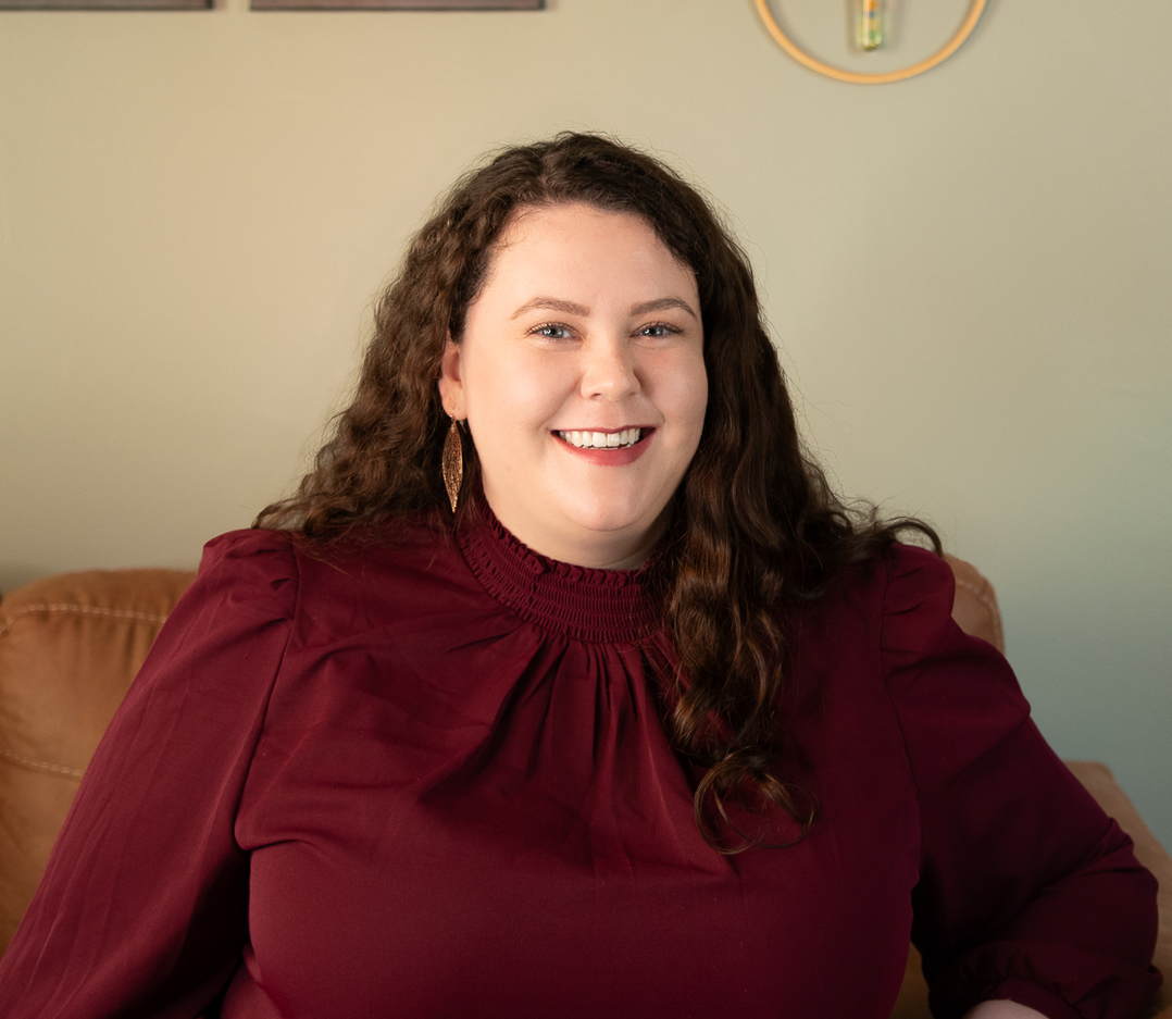 A woman in a maroon shirt is sitting on a couch and smiling.