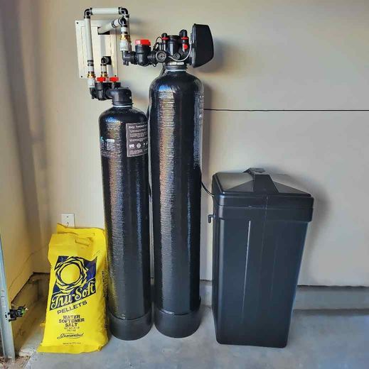Water softener system with two black tanks, a salt container, and a yellow bag of salt in a garage.