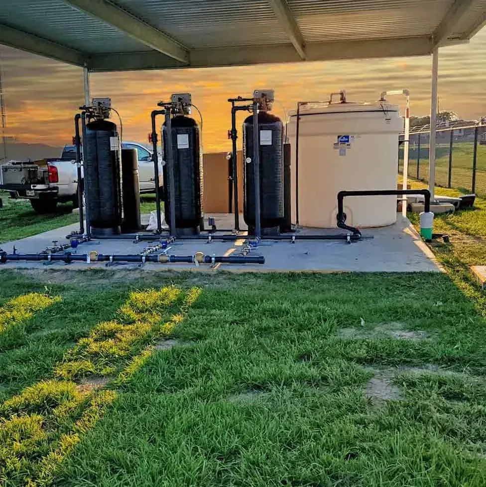 Water filtration system with tanks and pipes on concrete pad, green grass, and sunset.