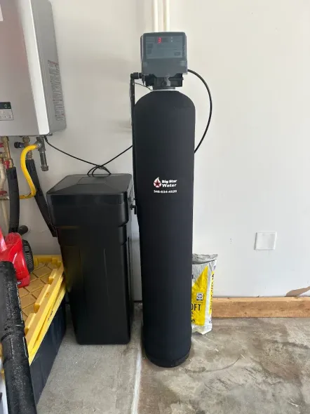 Water softener system in a garage: tall black tank, black salt container, and a grey water heater.