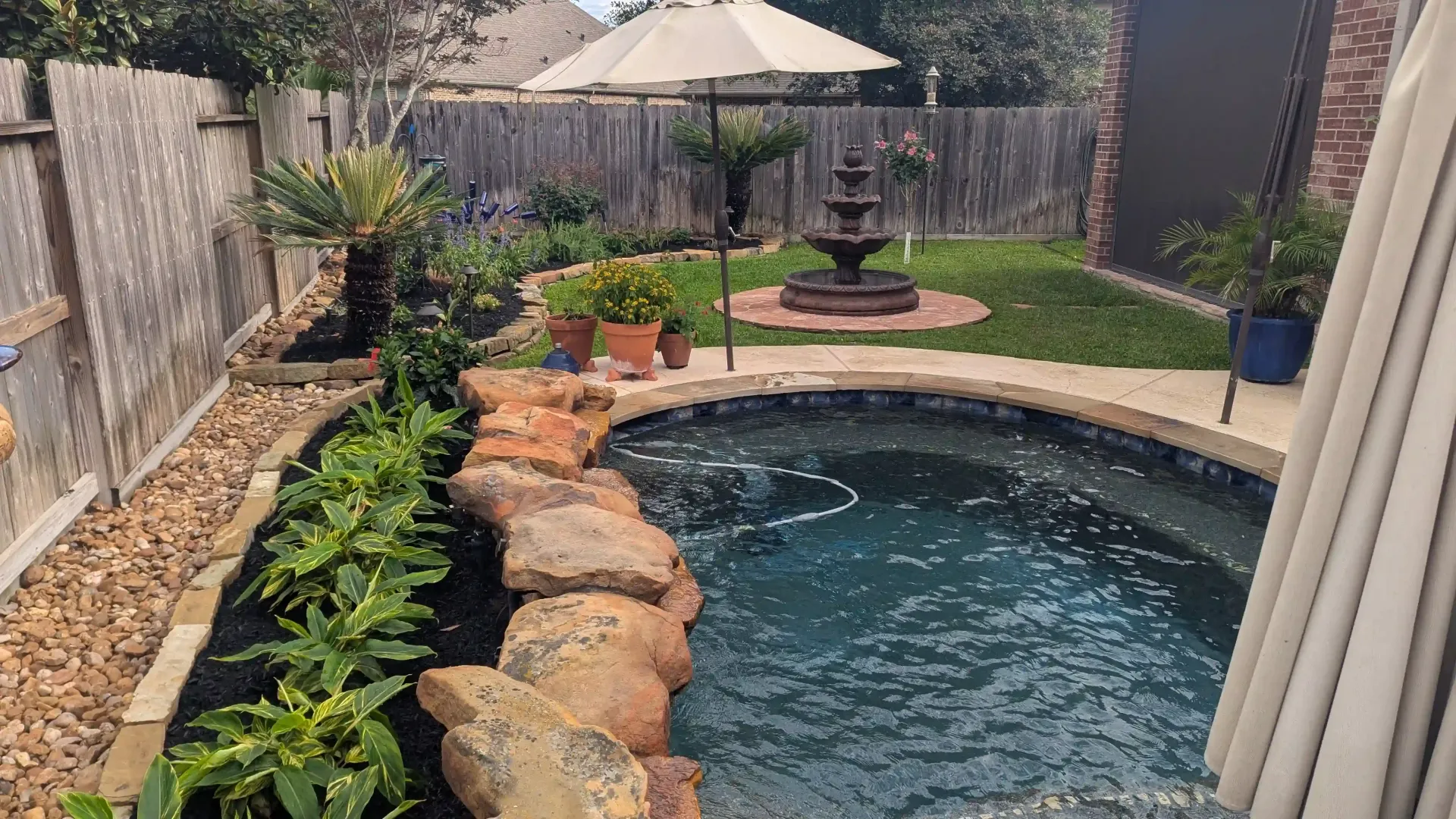 A small swimming pool in a backyard with a fountain and umbrella.