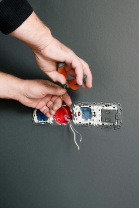 Hands wiring an electrical outlet with exposed red and blue wires on a gray wall