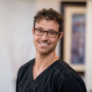 a man wearing glasses and a black scrub top smiles for the camera