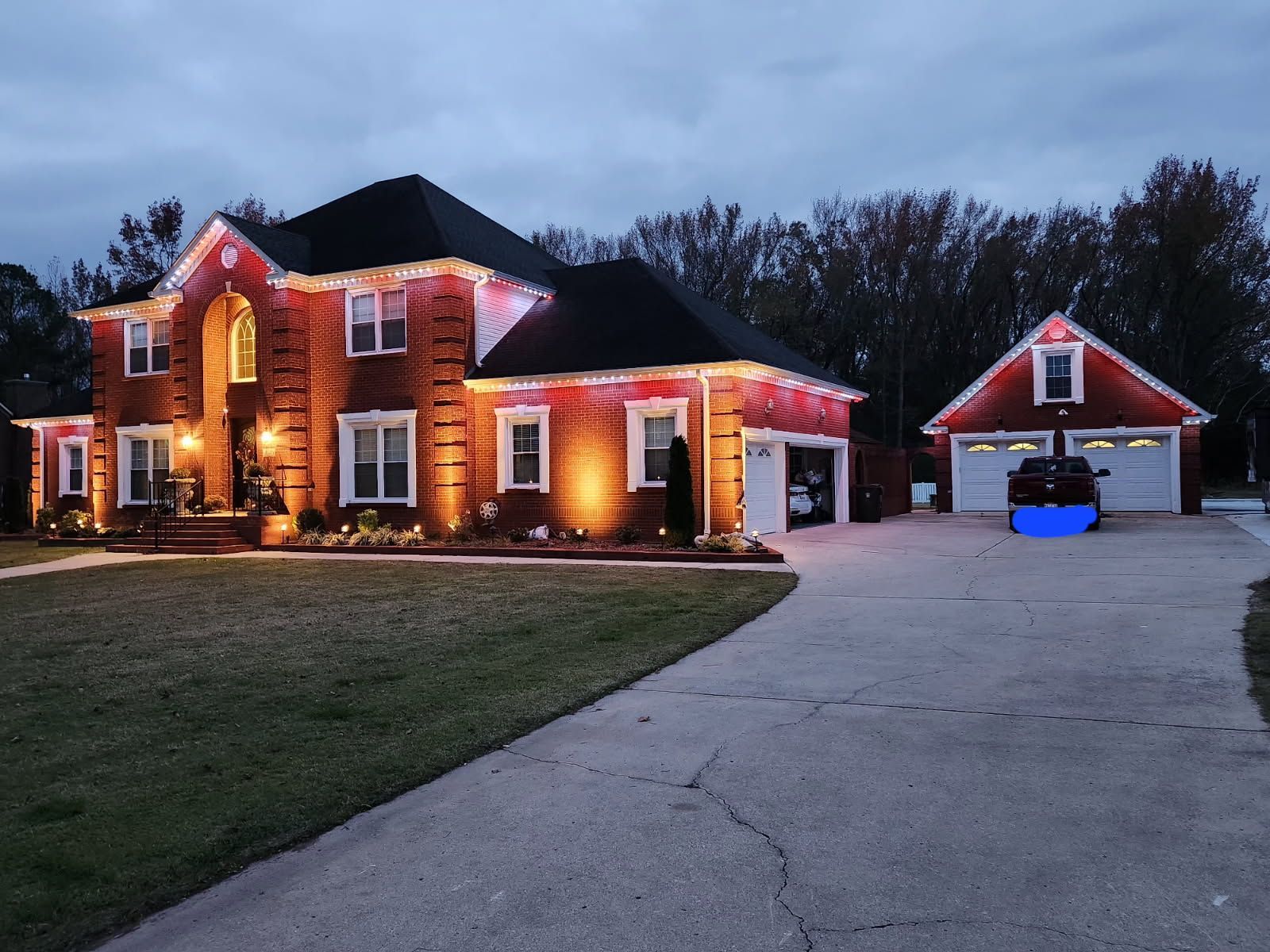 Brick house and detached garage illuminated at dusk, with lights outlining rooflines.