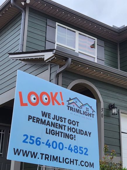Sign advertising permanent holiday lighting on a house with green siding, a window, and the Trimlight phone number.