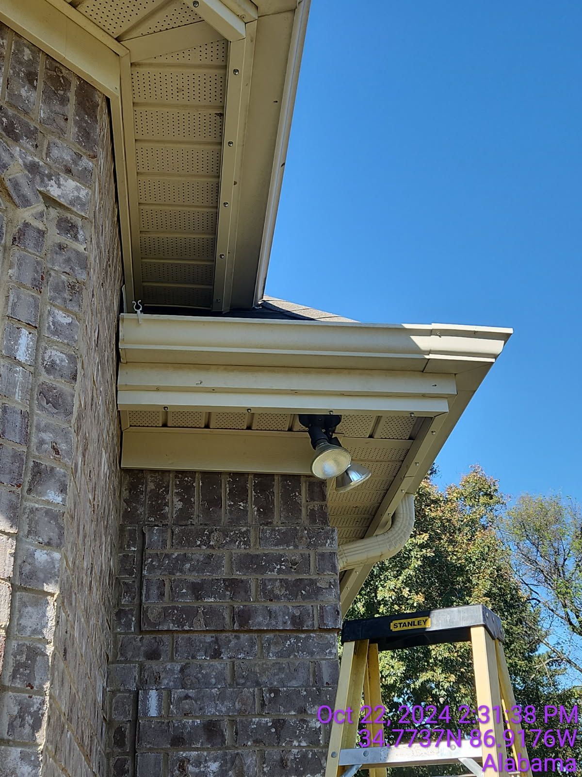 Brick building corner with tan trim, eaves, and a security light. Ladder propped nearby under blue sky.