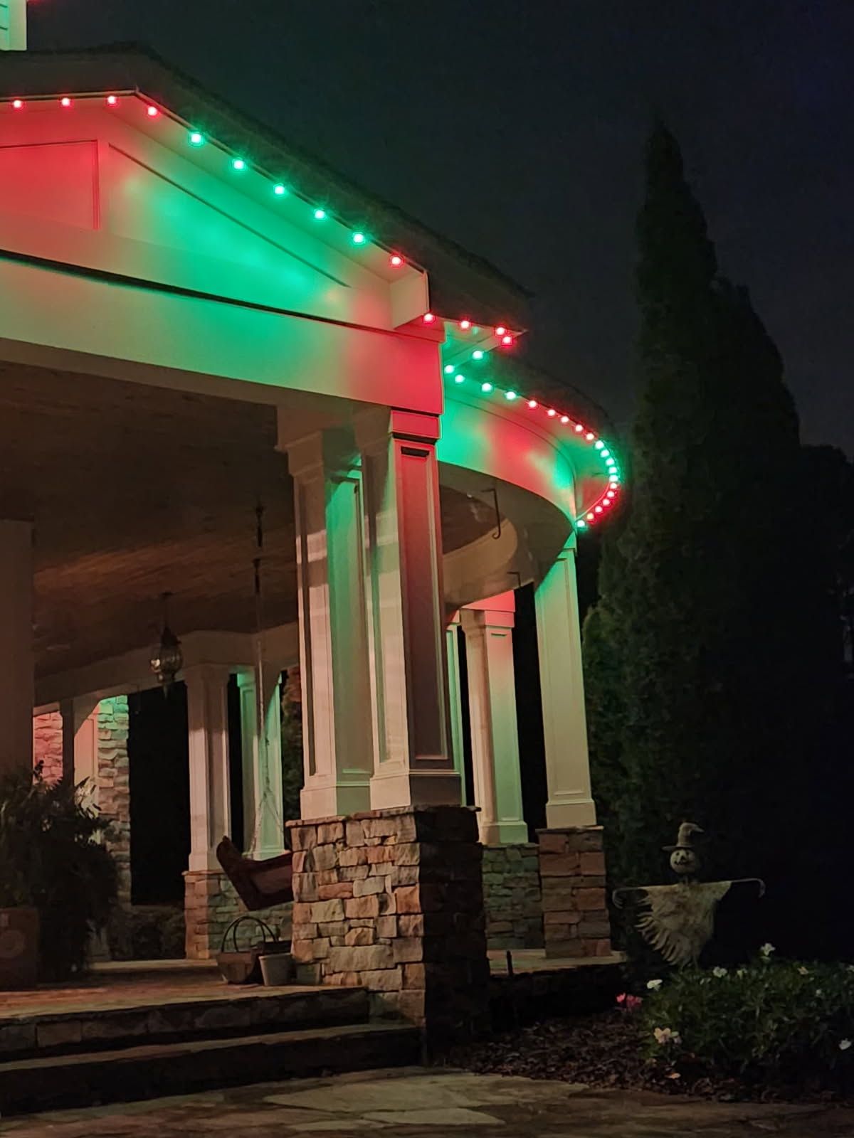 Christmas lights, red and green, illuminate a house's porch columns at night.