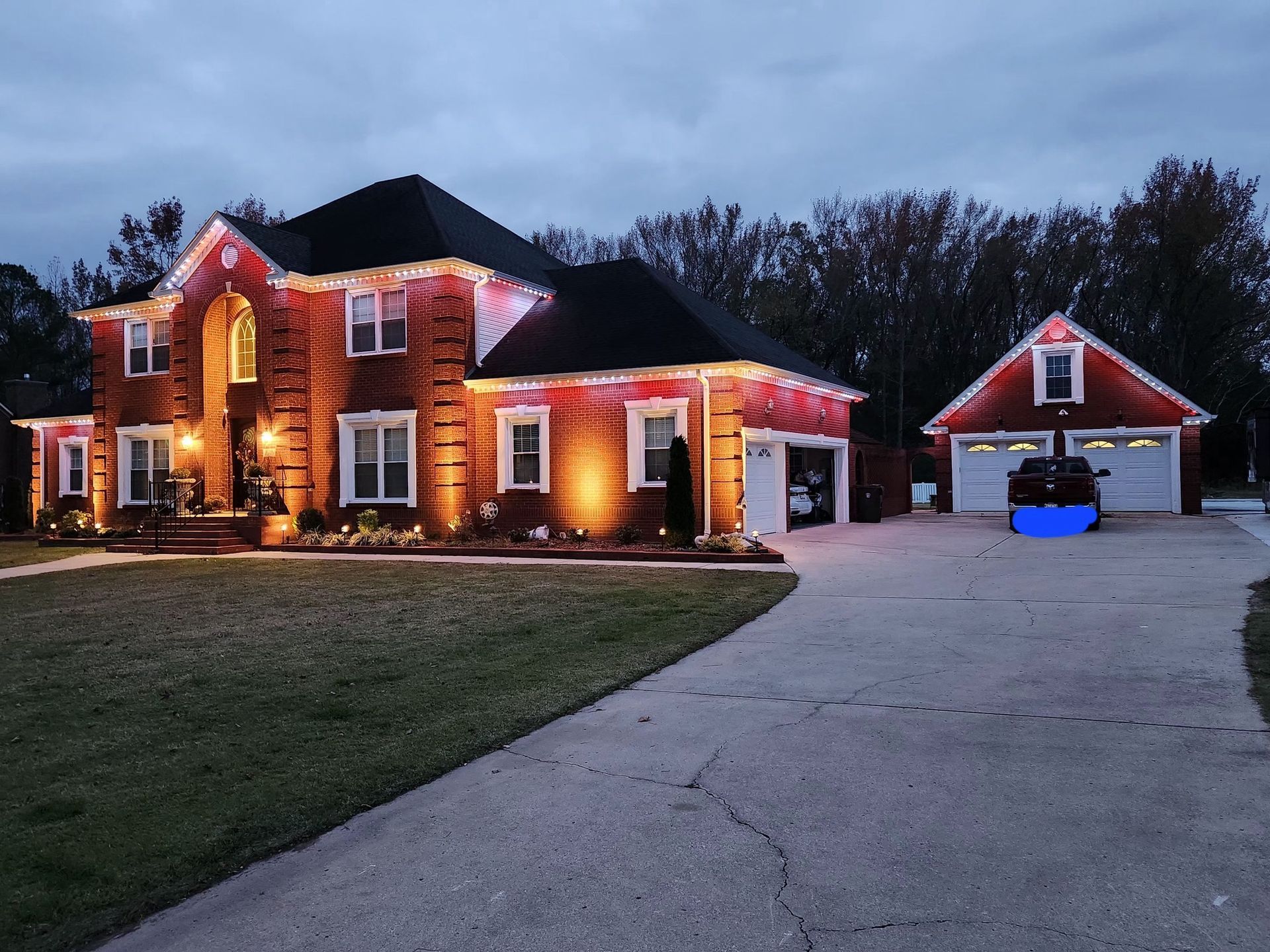 Brick house with Christmas lights, detached garage, and long driveway at dusk.