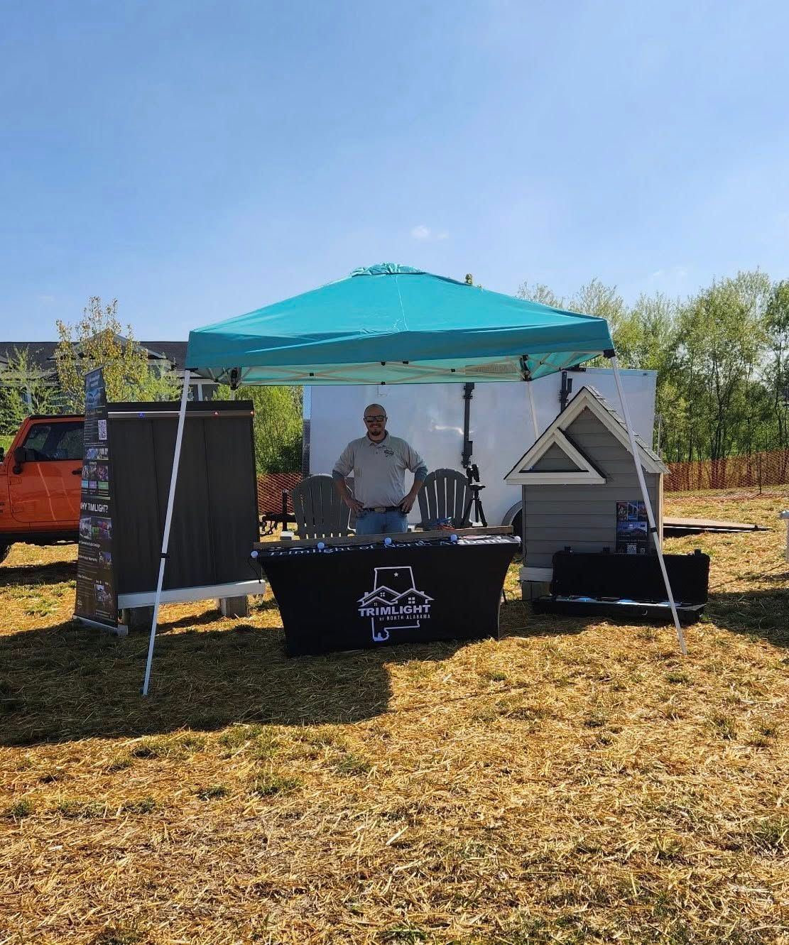 Man standing at a booth under a teal canopy, with dog house samples and display materials on a table, outdoors.