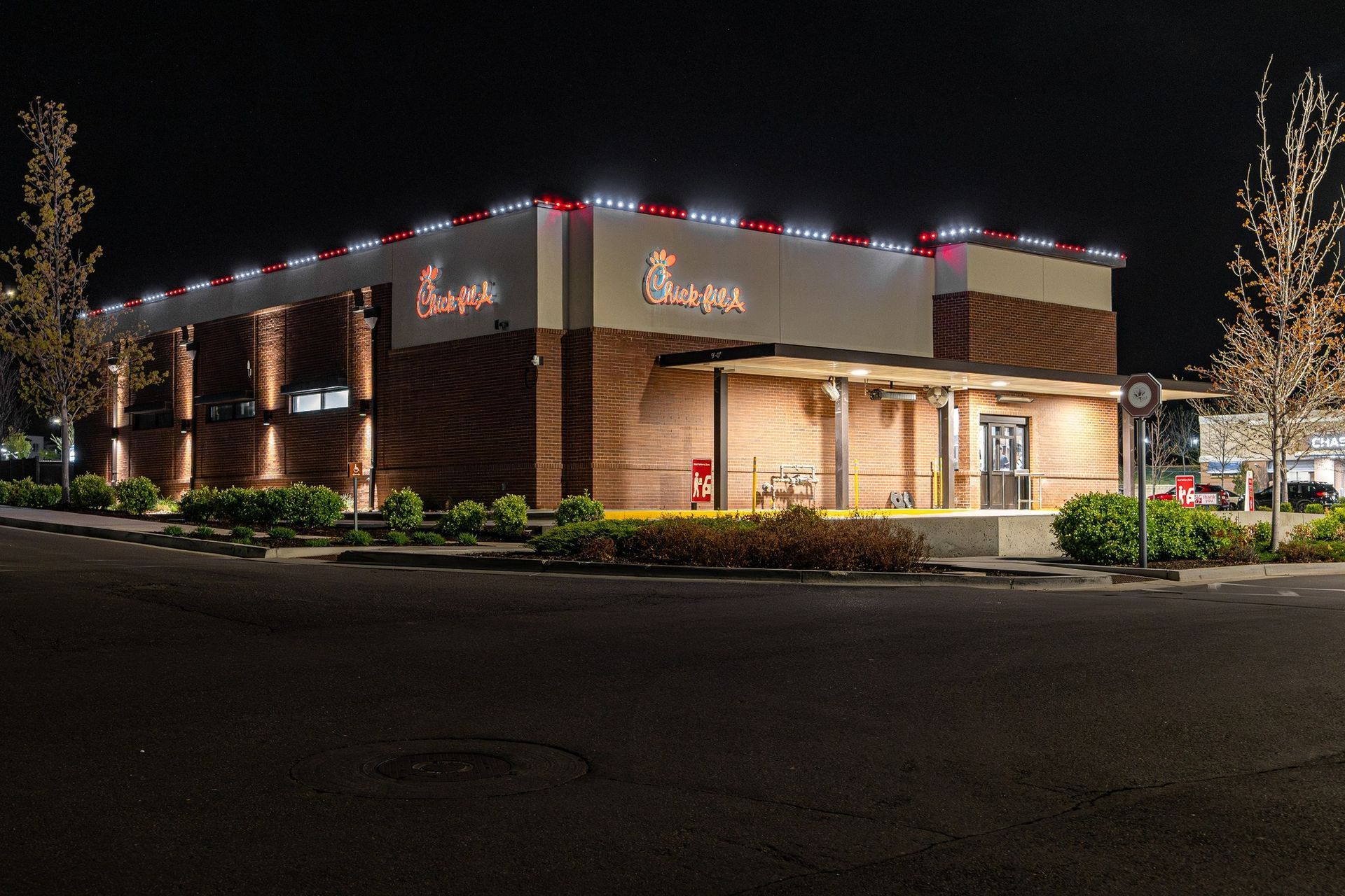 Chick-fil-A restaurant at night, exterior view. Building lit with red and white lights. Brick and neutral color siding.
