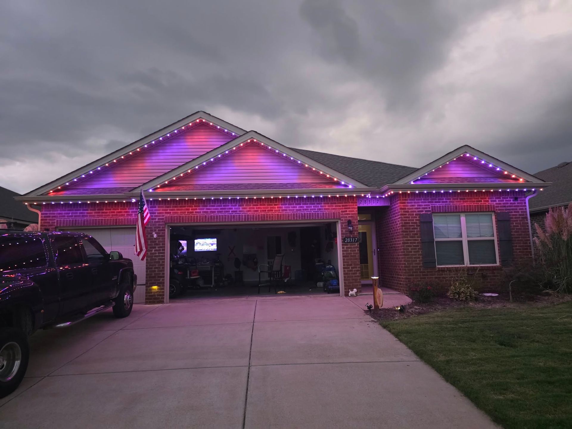 House with red and purple lights on roof and along the garage and window. Dark sky and truck parked in driveway.