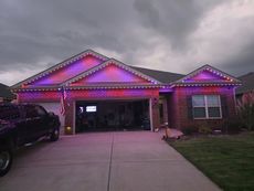 House with red and purple lights on roof and along the garage and window. Dark sky and truck parked in driveway.